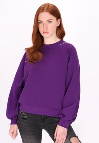 Lilla sweater med lange, puffede ærmer og ribbede manchetter. Har en rund halsudskæring og subtilt logo på skulderen.