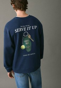 Námornícky modrý sveter s grafickým motívom kľúčenky a tenisovej loptičky na zadnej strane, s textom: "SERVE IT UP" a "GAME. SET. MATCH."