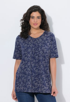 Donna con capelli ricci e scuri, indossa una maglietta a maniche corte blu con una leggera stampa floreale e jeans blu, in piedi contro uno sfondo bianco.