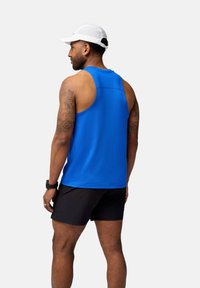 Blå sporttop med mesh-textur, kombinerad med svarta shorts. Modellen bär en vit keps och ett armbandsur.