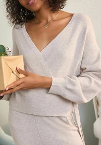 Svetlo bež ovit sweater s V-izrezom, rebrastimi manžetami in zavezanim detajlom ob strani. Model drži majhen zavit darilo z rastlino.