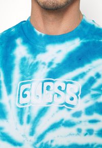 Sudadera azul con estampado tie-dye, con un cuello acanalado y un destacado logotipo "GUESS" en blanco, en un estilo elevado y bordado.