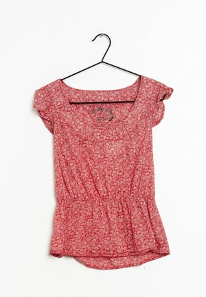 Red short-sleeve blouse with white floral pattern, gathered waist, and round neckline, hanging on a black metal hanger against a white background.