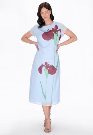 Donna in piedi con le mani sui fianchi, indossa un vestito midi azzurro chiaro con grandi stampe di fiori rossi e tacchi beige con cinturini.