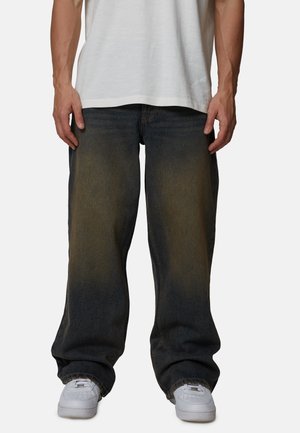 Man draagt losse donkere spijkerbroek met vervaagde plekken, witte sneakers en een wit T-shirt, staand tegen een effen achtergrond.