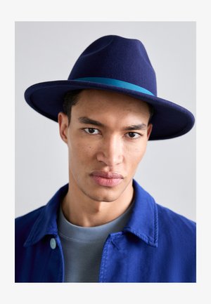 Navy felt hat with a wide brim and a light blue ribbon band. Model wears a blue jacket over a gray shirt, displaying a neutral background.