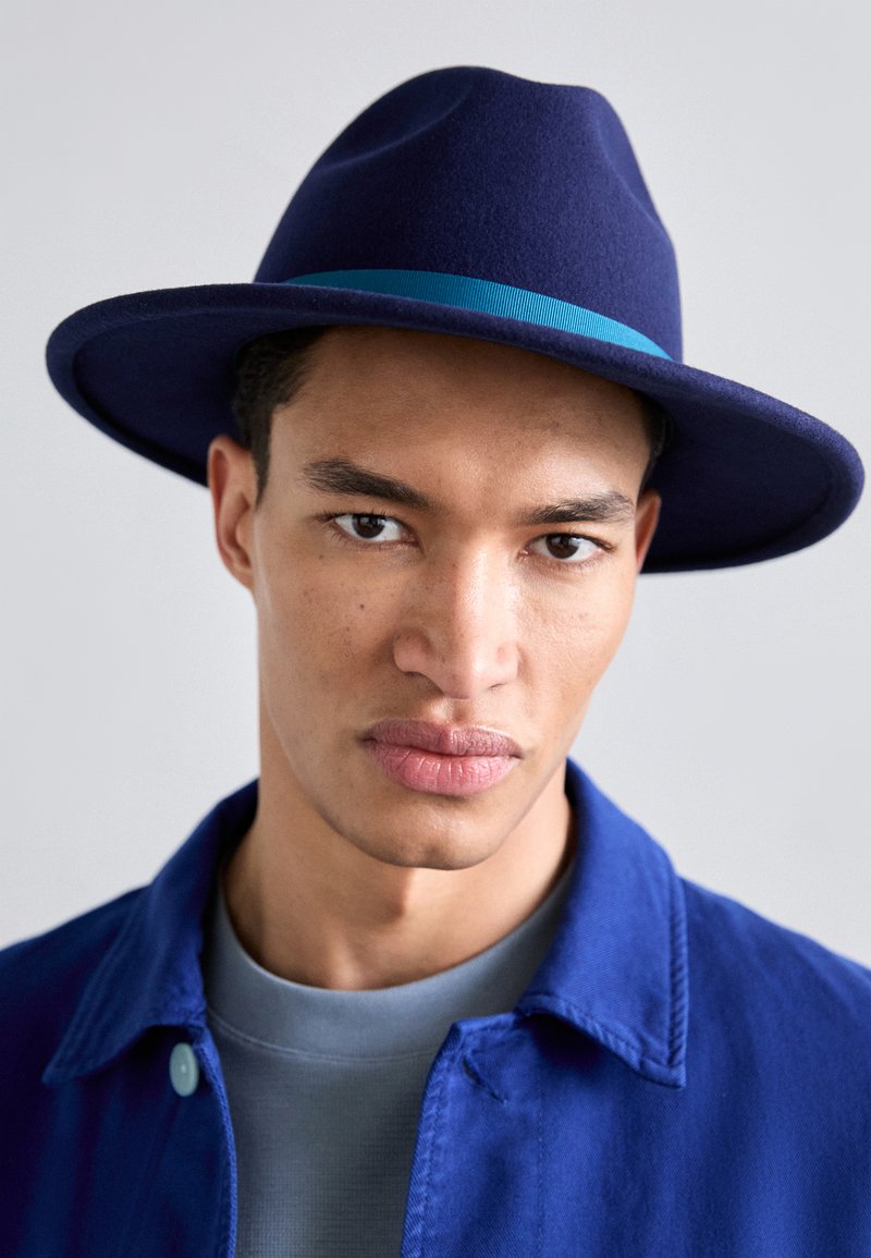 Navy felt hat with a wide brim and a light blue ribbon band. Model wears a blue jacket over a gray shirt, displaying a neutral background.