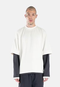 Biela oversized tričko s krátkymi rukávmi preložené cez tmavé tričko s dlhými rukávmi. Hladká textúra a okrúhly výstrih; neformálny dizajn.