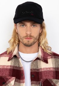 Gorra de tela negra con "Schott NYC" bordado en negro y rojo. Combinada con una camisa de franela a cuadros en rojo, blanco y gris.