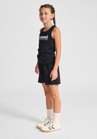 Svart idrottstopp med vit text "hummel PERFORMANCE", ihop med svarta shorts. Modellen har på sig vita skor med svarta accenter och ljusa strumpor.