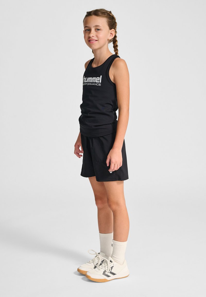 Svart idrottstopp med vit text "hummel PERFORMANCE", ihop med svarta shorts. Modellen har på sig vita skor med svarta accenter och ljusa strumpor.