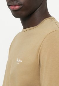 T-shirt marrone chiaro con scollo rotondo, caratterizzato da un logo bianco sul petto. Tessuto morbido con design aderente e dettagli di cucitura discreti.