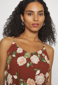 Top de tanque floral castanho com alças finas, apresentando grandes rosas em tons pastel e folhas verdes sobre um fundo de tecido texturizado. Brincos de argola prateados.