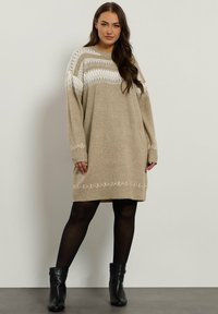 Robe pull en beige clair avec des motifs géométriques crème aux épaules et à l'ourlet ; poignets côtelés ; associée à des bottines noires.
