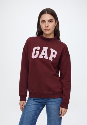 Kvinna med långt mörkt hår som bär en vinröd GAP-tröja och blå jeans, stående framför en enfärgad vit bakgrund.