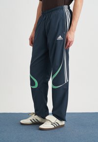 Tamsiai mėlyni sportiniai kelnės su žaliais akcentais ir baltais dryžiais, pagamintos iš lengvos medžiagos, su elastingu juosmeniu ir siaurėjančiomis kojomis.