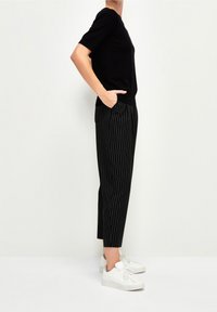 Černý top s krátkým rukávem ve spojení s černými kalhotami s proužky. Modelka má na sobě bílé tenisky. Oblečení má volný střih a hladkou texturu.