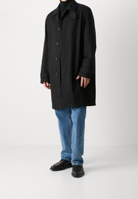 Abrigo negro impermeable con cuello y botones frontales, combinado con jeans de mezclilla azules y zapatos de cuero negros.