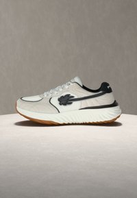 Άσπρο και μαύρο αθλητικό παπούτσι με καουτσούκ σόλα, δέσιμο με κορδόνια και μαύρο λογότυπο στο πλάι, τοποθετημένο σε ουδέτερη επιφάνεια και φόντο.