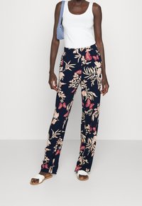 Mörkblå blommiga byxor med vida ben och rosa och krämfärgade botaniska mönster, kombinerade med en vit linne och sandaler.