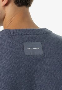 Tamsiai mėlyna megztinis su tekstūruotu paviršiumi ir raukinėtu apykakle, kuriame nugarinėje pusėje yra stačiakampė pilka etiketė su užrašu "PEGADOR".