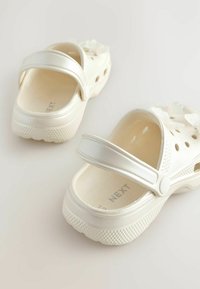Weiße Plastikschuhe mit glänzender Oberfläche, Fersenriemen, Ausschnitten für Belüftung und floralen Akzenten auf der Vorderseite. Texturierte Sohle für besseren Halt.