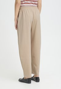 Pantalon beige à taille élastique avec une coupe ample, présentant une texture lisse et des jambes droites. Associé à des chaussures plates noires.