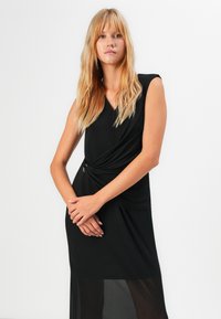 Kvinne med langt blondt hår som har på seg en ermeløs svart kjole med innsamlet midje, stående mot en enkel hvit bakgrunn.