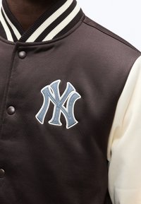 Κοντινό πλάνο προσώπου που φοράει καφέ και κρεμ varsity μπουφάν με ριγωτό γιακά και κεντημένο λογότυπο NY σε ανοιχτό μπλε στο στήθος.