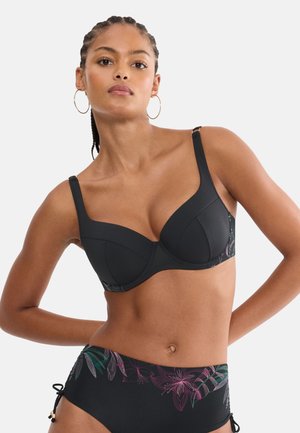 Frau trägt schwarzen Bikini mit lila und grünen Blumenmustern, posiert mit den Händen hinter dem Rücken vor einem einfachen hellen Hintergrund.