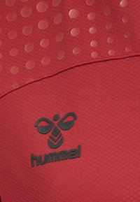 Röd sporttop med en texturerad tyg med upphöjda prickar och en svart logotyp av en stiliserad bi ovanför varumärket "hummel."