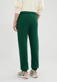 Zielone dresowe spodnie wykonane z gładkiego materiału, wyposażone w elastyczny pasek w talii oraz ściągacze na kostkach, noszone z beżowymi sneakersami i wzorzystymi skarpetkami.