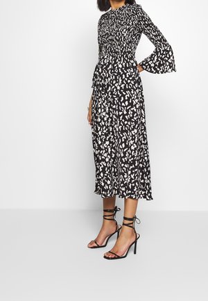 Frau trägt ein schwarz-weiß gemustertes Midikleid mit langen Glockenärmeln und schwarzen Knöchel-bindenden High Heel Sandalen.