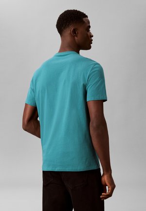 Άνδρας με την πλάτη, φοράει μονόχρωμο πετρόλ κοντομάνικο μπλουζάκι και σκούρο παντελόνι σε ουδέτερο γκρι φόντο.