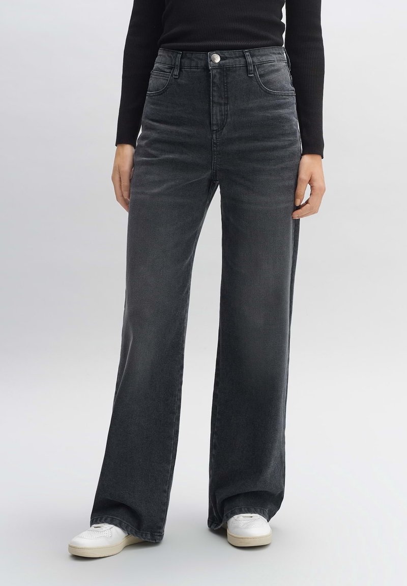 Personne portant un jean gris foncé à taille haute et jambes larges, un haut noir à manches longues, et des baskets blanches, debout sur un sol gris.