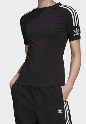 Osoba ubrana w czarny sportowy strój Adidas z białymi paskami na rękawach i spodniach, ręce w kieszeniach, widoczny tułów, jednolite tło.