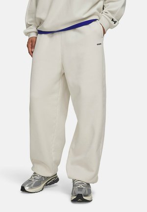 Person trägt lockere, off-white Sweatpants mit Seitentaschen, kombiniert mit grauen und schwarzen Laufschuhen und einem beigen Sweatshirt mit Logo auf dem Ärmel.