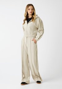 Beige hoodie met rits en zwarte strepen langs de mouwen, gecombineerd met hoge taille, wijde broek. Soepele textuur, casual pasvorm.