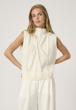 Maglione senza maniche bianco con un collo alto, caratterizzato da motivi a trecce testurizzati e orlo a coste, abbinato a pantaloni di colore chiaro.