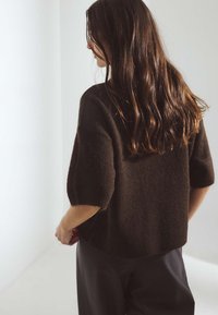 Dunkelbrauner, kurzärmeliger Pullover mit strukturiertem Finish und geripptem Saum, kombiniert mit schwarzen Hosen. Das Model ist von hinten zu sehen.