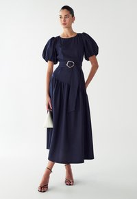 Navyblauwe jurk met pofmouwen, een taillering met een riem met een gouden gesp, en een vloeiende rok. Gecombineerd met sandaaltjes met banden en een witte tas.