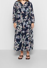Květinové maxi šaty v námořnické modři s dlouhými rukávy, výstřihem do V a nařaseným pasem. Obsahují patrovou sukni a barevný botanický potisk. Hnědé sandály.