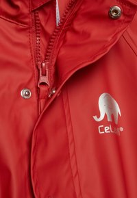 Červená voděodolná bunda s texturovaným povrchem, se zapínáním na přední straně a stříbrným logem slona, spolu s detailem na patentce.