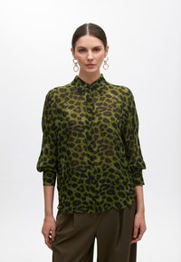 Zelená priesvitná košeľa na gombíky s čiernym vzorom leopardy, má dlhé rukávy a klasický golier.