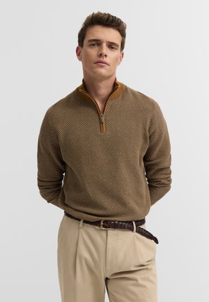 Junger Mann steht mit den Händen hinter dem Rücken, trägt einen braunen gemusterten Reißverschluss-Pullover und beige Hosen mit geflochtenem Gürtel.