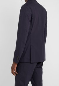 Marinblazer i lättviktsmaterial, enkel vent i ryggen, tre knappar på varje ärm, skräddarsydd passform, rena linjer, minimal detaljering.