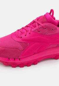 Chaussure de sport rose vif en textile synthétique et suède, avec une empeigne texturée, un col rembourré et une semelle en caoutchouc épaisse avec adhérence.