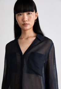 Camisa azul marinho transparente com decote em V, colarinho curto e dois bolsos no peito. Tecido leve com uma textura suave e brilho subtil.