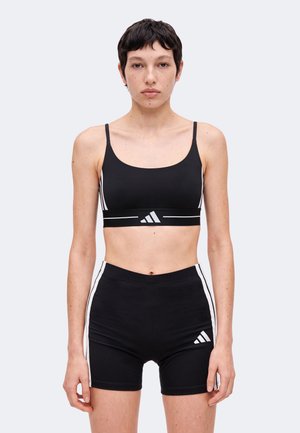 Jeune personne avec cheveux courts et foncés portant un brassière de sport noire Adidas et un short noir assorti avec des bandes blanches, debout devant un fond clair.