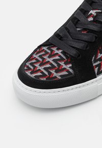 Bout de basket noir avec lacets noirs, semelle blanche et tissu à motif en zigzag géométrique noir, blanc et rouge.